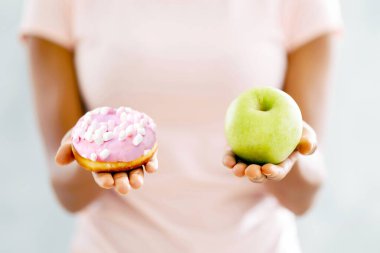 Sağlıklı mı abur cubur mu? Açık arkaplanda çörek ve elma arasında seçim yapan siyahi kadının yakın görüntüsü. Milenyum hanımefendisi yiyecekleri hakkında karar veriyor, baştan çıkarılmayla yüzleşiyor.