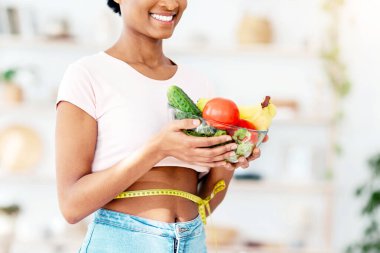 Kilo verme konsepti için sağlıklı beslenme. Elinde mezura ile evde taze meyve ve sebze dolu bir kaseyle mutlu siyah bayan, yakın plan. Milenyum Afrikalı Amerikalı kadın vegan diyetinin tadını çıkarıyor