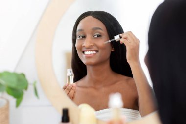 Rutin vücut güzellik hizmetleri, sabah prosedürleri ve evdeki yalnızlık sırasında tedavi. Bir şişe nemlendirici serumla gülümseyen Afro-Amerikan kadın, pipetle cildine ürün uyguluyor.