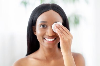 Binlerce yıllık neşeli Afro-Amerikan bayan aynaya bakar, pamuk pedi ve kozmetik ürünleriyle deriyi temizler, yatak odasının iç kısımlarındaki duştan sonra sabah bakım rutininin keyfini çıkarır.