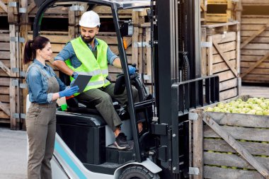 Meyve suyu ve püre üretimi, depoda çalışmak, yükleme için modern cihazlar. Mutlu genç kadın tabletini forkliftte yeşil elmalı sert şapkalı adama gösteriyor, ahşap kutularda.