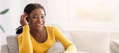 Afro-Amerikan kadın iç mekanda poz veriyor, rahat koltukta oturuyor, yastığa sarılıyor ve kameraya gülümsüyor, siyah kadın oturma odasında dinleniyor, boş vakitlerin tadını çıkarıyor, boşluğu kopyalıyor.