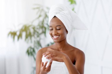 Kırışıklıklara ve deri sorunlarına karşı kozmetik ilaçlar. Duş aldıktan sonra beyaz havlular içinde genç, mutlu, Afro-Amerikan kadın blogcu. Minimum yatak odasının içinde, boş bir yerde krema kavanozu açıyor.