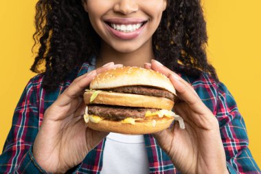 Abur cubur. Biftek ve peynirli hamburger tutan komik, Afro-Amerikan kadının yakın plan portresi. Kıvırcık saçlı, lezzetli sandviçi yemeye hazır, turuncu arka planda izole edilmiş siyah kadın, afiş.