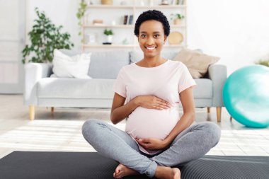 Hamilelik sırasında formda kalmak. Güzel, siyahi, hamile bir kadın karnına dokunuyor, evde spor minderinin üzerinde oturuyor. Müstakbel anne spor yaparken kameraya bakıp gülümsüyor.
