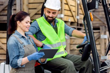 Akıllı depo yönetim sistemi. Kasklı milenyum sakallı adam forklift kullanıyor. Tabletli koruyucu eldivenli gülen kadın yöneticiyle sırayı tartışıyor. Bir sürü tahta kutu var.