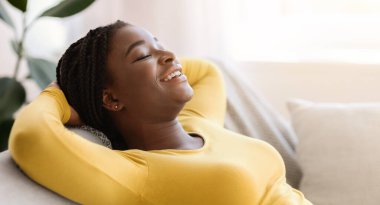 Yurtiçi rahatlama. Gülümseyen siyah bir kadının portresi arkası kapalı gözler ve ellerle oturma odasında rahat bir kanepeye uzanmış, boş bir manzarası ve boş bir yeri olan,