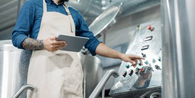 Bira fabrikasında çalışan erkek işçi, yönetim ve mobil uygulama. Meşgul erkek çalışan veya giysi sahibi dijital tabletle bira fabrikasında ekipmanı kontrol ediyor ve düğmeye basıyor, kırpılıyor, boş yer, panorama
