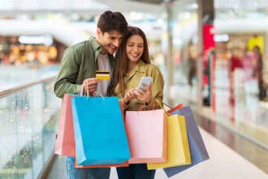 Mobile Offer. Happy Couple Shopping Online With Cellphone And Credit Card Using Shopper Application Standing With Colorful Bags In Modern Mall On Weekend. Ecommerce Concept