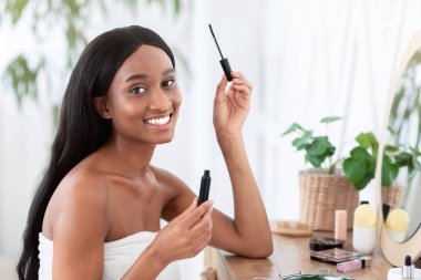 Sabah makyajı. Milenyum Afrikalı Amerikalı bir kadın kirpiklerine rimel sürüyor ve kameraya bakıyor, kozmetik ürünleriyle masaya oturuyor, sabahları modern yatak odasının içinde, uzay fotokopisi çekiyor.