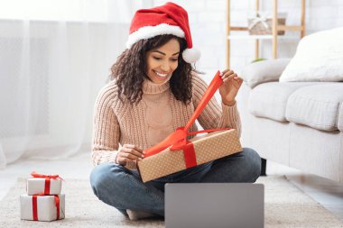 Heyecanlı siyah kadın Noel hediyesiyle oturup hediyesini açıyor oturma odasında dizüstü bilgisayarla kırmızı kurdeleli bir hediye kutusunu açıyor, internette kutlama yapıyor, evde oturuyor ve Noel 'i kutluyor.