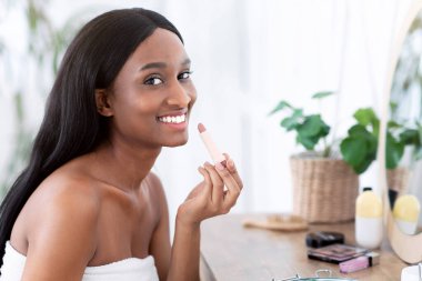 Karantina sırasında evde dudak bakımı, gündüz makyajı ve güzellik bakımı. Bin yıllık neşeli Afro-Amerikan kadın havluyla masaya oturuyor ve sabah yatak odasına ruj sürüyor, içi boş.