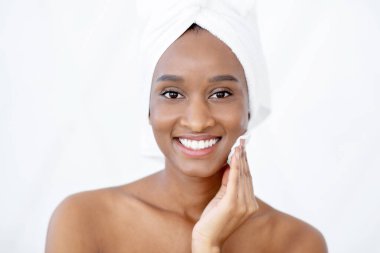 Güzellik bakımı, makyajı sil ve evde tonik kullan. Beyaz havluyla çekici genç Afrikalı Amerikalı bayan beyaz arka planda duş aldıktan sonra yüzüne pamuk ped ve tonik sürterek boşluğu kopyaladı.