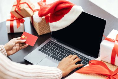 Noel için hazırlanıyorum. Kredi kartı tutan Afrikalı Amerikalı kadının omuz hizasında ve ekranda siyah beyaz ekranlı bilgisayar kullanıyor ve Noel Baba şapkası takıyor.