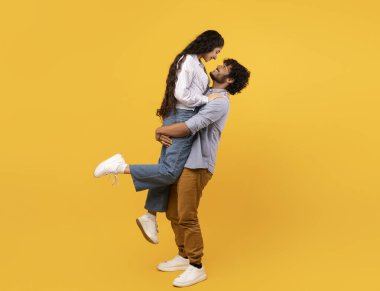 Loving indian guy lifting his girlfriend and holding her on yellow studio background, profile side view, free space banner. Romantic couple expressing their love to each other