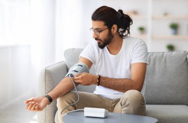 Evdeki kanepede oturan genç Hintli adam kan basıncını ölçüyor. Orta Doğulu beyaz tişörtlü adam modern tonometre kullanıyor. Stres kaynaklı hipertansiyon yaşıyor.