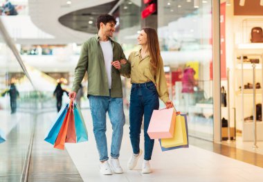 Aile alışverişi. Renkli alışveriş torbaları taşıyan gülümseyen bir çift. Büyük satışlar sırasında el ele tutuşup modern hipermarkette yeni kıyafetler alıyorlar. AVM Konseptinde Memnun Müşteriler