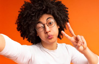 Zamanı kandırmak. Bin yıllık adam komik selfie yapma, barış işareti gesturing, göz kırpma ve dudaklar üfleme, turuncu arka plan