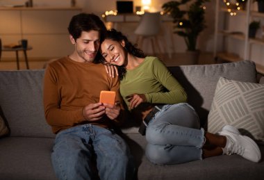 Genç çift cep telefonu kullanıyor. Hafta sonları akşamları evdeki koltukta oturuyorlar. Millennial Koca ve Eş Akıllı Telefon Konsepti ile İnternet Taraması