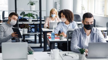 Strateji ve pazarlama planı oluşturuluyor. Koruyucu maskeli genç adam, iş yerindeki Afrikalı Amerikalı meslektaşına bilgisayarlı antiseptikle iş yerinde tablet gösteriyor.