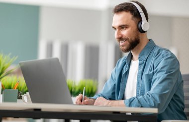 Çevrimiçi Eğitim. Kablosuz kulaklıklar takan ve not alan gülümseyen genç adam, Milenyum erkeği Webinar 'ı izliyor, İnternet dersine katılıyor, ofisteki masasında oturuyor.