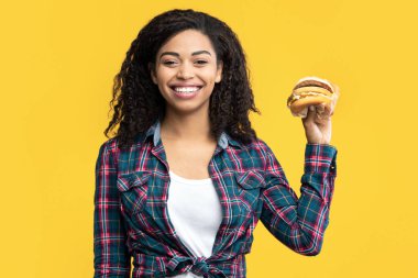 Fast Food aşığı. Kıvırcık saçlı, gülümseyen Afro-Amerikan kadının portresi büyük lezzetli bir burger tutuyor ve kameraya gösteriyor. Siyah Kadın Sandviç Teklif Ediyor, Turuncu Arkaplan, Bayrak