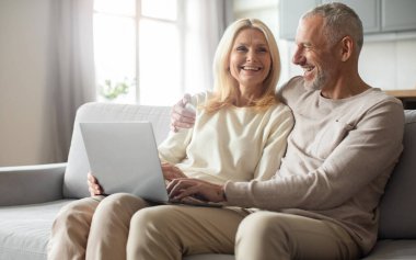 Neşeli bir son sınıf çifti, güneşli bir oturma odasındaki rahat kanepelerinde dizüstü bilgisayar kullanırken kahkahalarını paylaşıyor. Rahat ve samimi zamanları güçlü bağlarını vurguluyor..