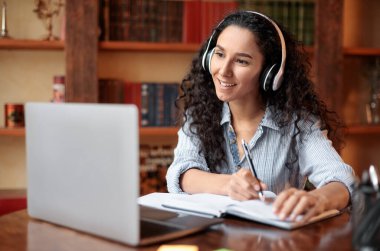 Modern Teknoloji ve Eğitim. Kablosuz kulaklıkla gülümseyen kadın Webinar izliyor, masada oturuyor, dizüstü bilgisayar kullanıyor ve not defteri yazıyor, not alıyor. Karantina Sırasındaki Eğitim