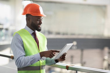 Güvenlik yeleği ve miğfer mühendisi ya da mimar olan gülümseyen Afro-Amerikan genç adamın yan görünüşü dijital tabletle inşaat alanını kontrol ediyor, modern teknolojiler kullanıyor, uzay kopyaları yapıyor.