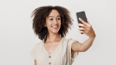 Modern blogcu sosyal ağlar veya bloglar için fotoğraf çekiyor. Kıvırcık saçlı, mutlu Afrikalı Amerikalı kadın öğrenci selfie çekiyor, arka planda izole edilmiş, stüdyo çekimi, boş alan.