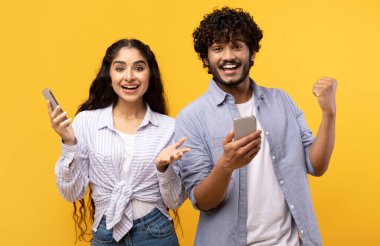 Online piyango. Heyecanlı genç Hintli erkek ve bayan sevinçle seviniyor ve ellerinde akıllı telefonlar, sarı arka planda durup zaferi haykırıyorlar.