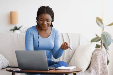 Kulaklıklı Milenyum Afrikalı Kadın evde dizüstü bilgisayarda giyiniyor, çevrimiçi eğitim alıyor, bilgisayarla çalışıyor ve notlar alıyor, uzaktan öğrenmenin keyfini çıkarıyor, boşluğu kopyalıyor.