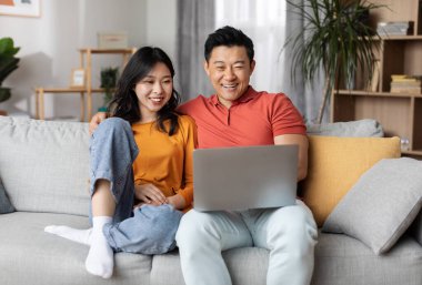 Sevgi dolu Çinli çift birlikte dizüstü bilgisayar kullanıyor, ev içinde rahat, mutlu Asyalı adam güzel karısına sarılıyor ve defter ekranına bakıyor, fotoğraflar izliyor, uzay kopyaları kopyalıyor.