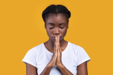 Barışçıl Afro-Amerikan Genç bayan dua eden ellerini sıkıca tutarak dua ediyor. Sarı arka plan üzerindeki stüdyoda gözleri kapalı poz veriyor. Tanrı 'ya Dua Gönderen Siyah Kadın Portresi
