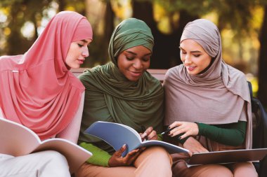 Şehir parkının dışında oturmuş kitap okuyan üç mutlu Müslüman öğrenci. Arap Kadınları için Üniversite Diploması, Üniversite Eğitim Yaşam Tarzı Konsepti.