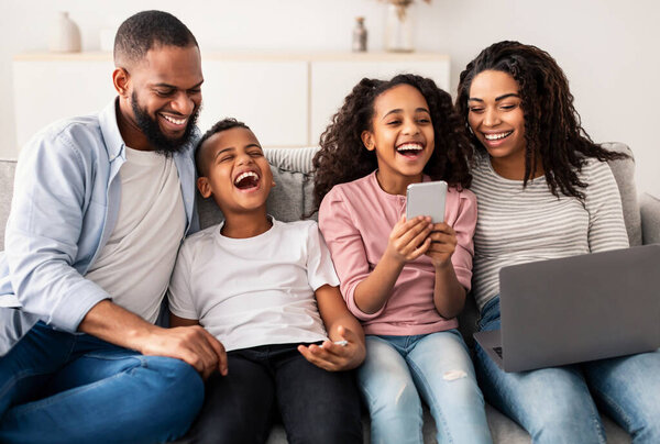 Entertainment And Leisure. Portrait of happy African American parents and their little children using laptop and smartphones, spending time together sitting on the couch in living room and laughing
