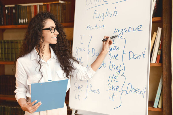 Teacher In Classroom. Smiling professional female English teacher writing on whiteboard with marker. Young woman wearing eyeglasses, explaining grammar rules to the students, standing with folder