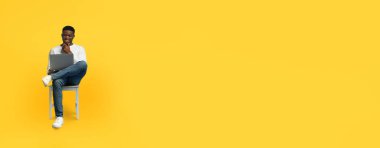 Genç bir Afrikalı Amerikalı adam bir sandalyede oturuyor, dizüstü bilgisayar kullanırken gülümsüyor. Günlük giysiler giyer ve iş ya da çalışmayla meşguldür. Etrafı parlak sarı bir zemin ile çevrilidir..
