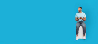 Açık mavi gömlekli genç bir Afro-Amerikalı beyaz bir taburede oturuyor, gülümsüyor ve telefonuna bakıyor. Canlı mavi arka plan neşeli atmosferi güçlendirir..