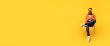 Sakallı bir adam bir sandalyeye oturmuş, basılı bir materyali okumakla meşgul. Canlı sarı arka plan, onun rahat duruş ve ifadesine odaklanmasını sağlar..