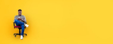 Afro-Amerikalı genç adam siyah ve kırmızı bir sandalyede rahat bir şekilde oturuyor, akıllı telefonuyla canlı sarı bir zemin üzerinde geziniyor. Bu aydınlık ortamda rahat ve memnun görünüyor..