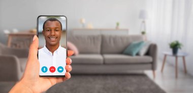 Bir kişi video görüşmesinde gülümseyen bir kişiyi gösteren bir akıllı telefon tutuyor. Dekorasyon, rahat bir oturma odası. Kanepe ve ev dekorasyonu var. İletişim için mükemmel..