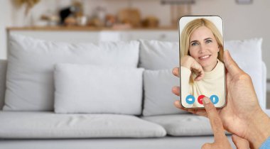 Evde bir kişi, gülümseyen yaşlı bir kadınla akıllı telefondan iletişim kurarken çevrimiçi bir video görüşmesi yapıyor. Rahatlatıcı ayarlar arka planda rahat bir kanepe ile geliştirilir..