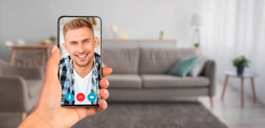 Bir kişi video görüşmesi sırasında gülümseyen bir yüz gösteren bir akıllı telefon tutuyor. Konforlu oturma odasında bir kanepe ve dekoratif öğeler var. İletişim için hoş bir ortam yaratıyor..