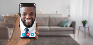 Bir kişi video görüşmesi sırasında gülümseyen bir kişiyi gösteren bir akıllı telefon tutuyor. Arka planda kanepesi ve bitkileri olan rahat bir oturma odası var. Sıcak bir atmosfer yaratıyor..