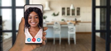 Bir kişi akıllı telefon kullanarak bir video görüşmesine katılıyor. Arka planda rahat ve neşeli bir ev ortamını gösteren şık bir mutfak var..