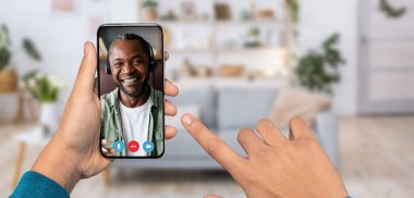 Birisi video görüşmesi için akıllı telefon kullanıyor, diğer uçta gülümseyen Afrikalı Amerikalı adam var. Arka plan, çiçekli ve kanepeli rahat bir ev ortamını gösteriyor..