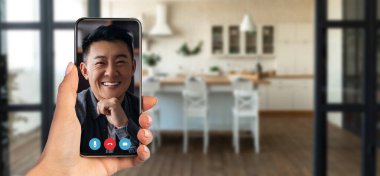Bir kişi video görüşmesinde akıllı telefon kullanarak konuşuyor. Arka planda ahşap zeminli ve temiz, rahat bir atmosferi olan modern bir mutfak var..