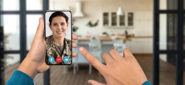 Bir kişi bir video araması yoluyla bir arkadaşla bağlantı kurmak için akıllı telefon kullanıyor. Ev dekorunda modern bir mutfak var ve odak noktası iletişim ve bağlantı..