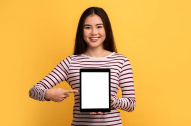 Uzun saçlı Asyalı genç bir kadın, elinde tuttuğu boş bir tableti işaret ederken coşkuyla gülümseyen canlı sarı bir fon karşısında duruyor. Çizgili gömleği neşe katıyor.